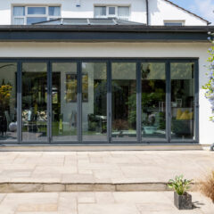 Modern house with large glass folding doors opening to a stone patio, surrounded by lush greenery and flowering bushes on a sunny day.