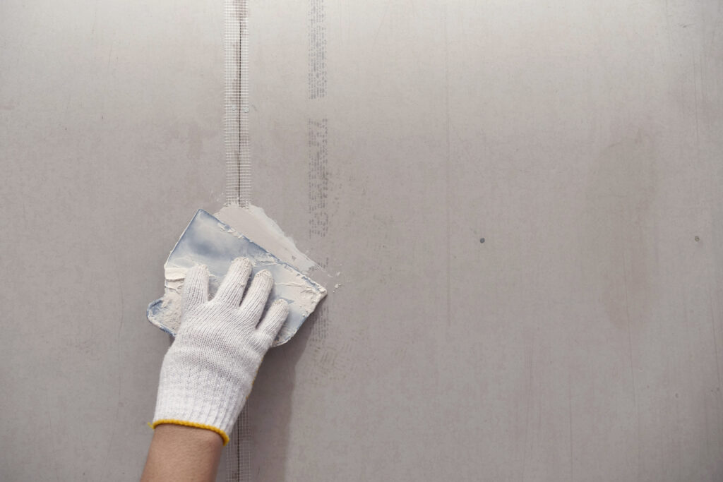 A gloved hand uses a putty knife to apply joint compound to a seam on a drywall panel, smoothing the surface for finishing.