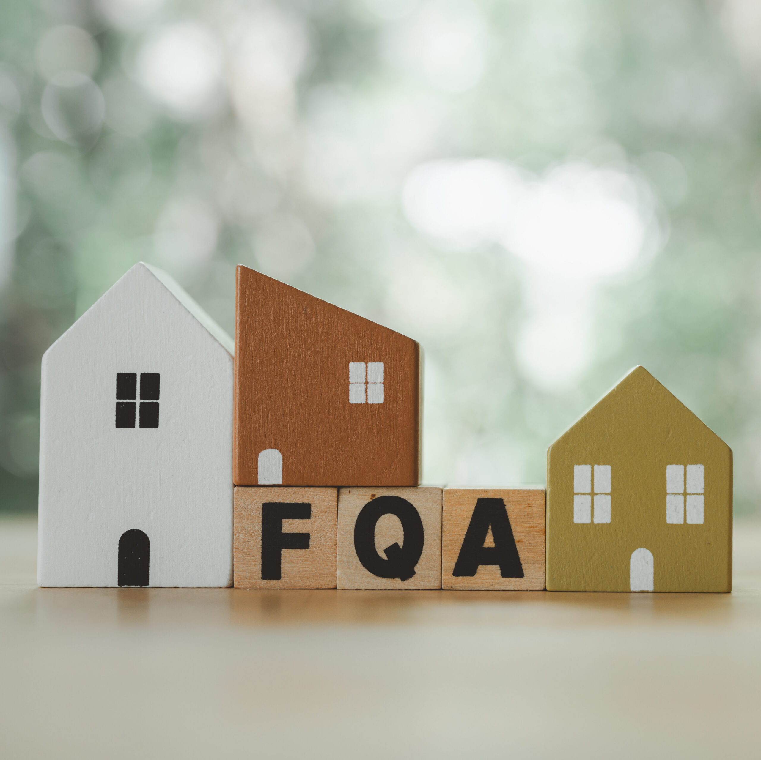 Three small wooden house models in white, brown, and green stand behind wooden blocks spelling "FAQ", with a blurred green and white background.